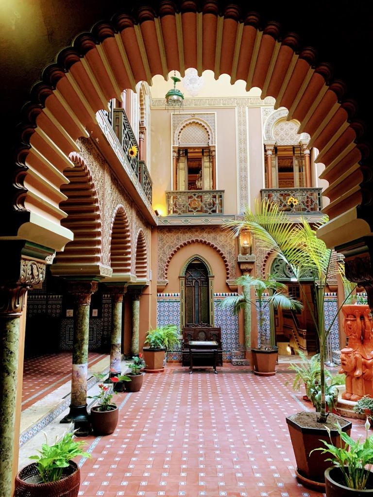 An islamic style courtyard with lush plants, arched doorways and blue and red tiles