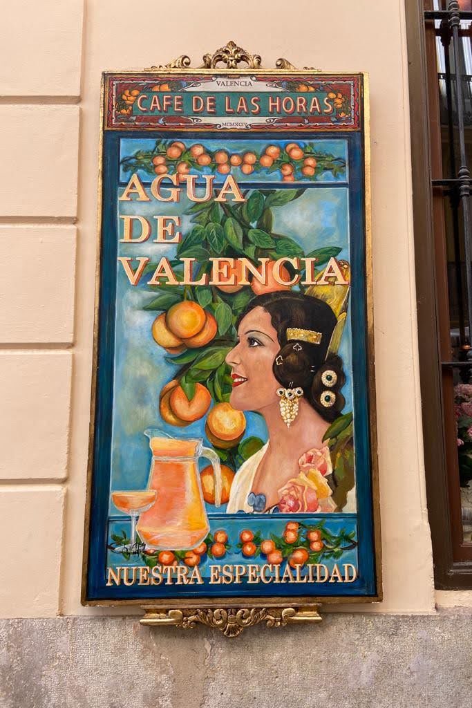 A painted sign of a woman looking at an orange tree that says "Agua de Valencia"