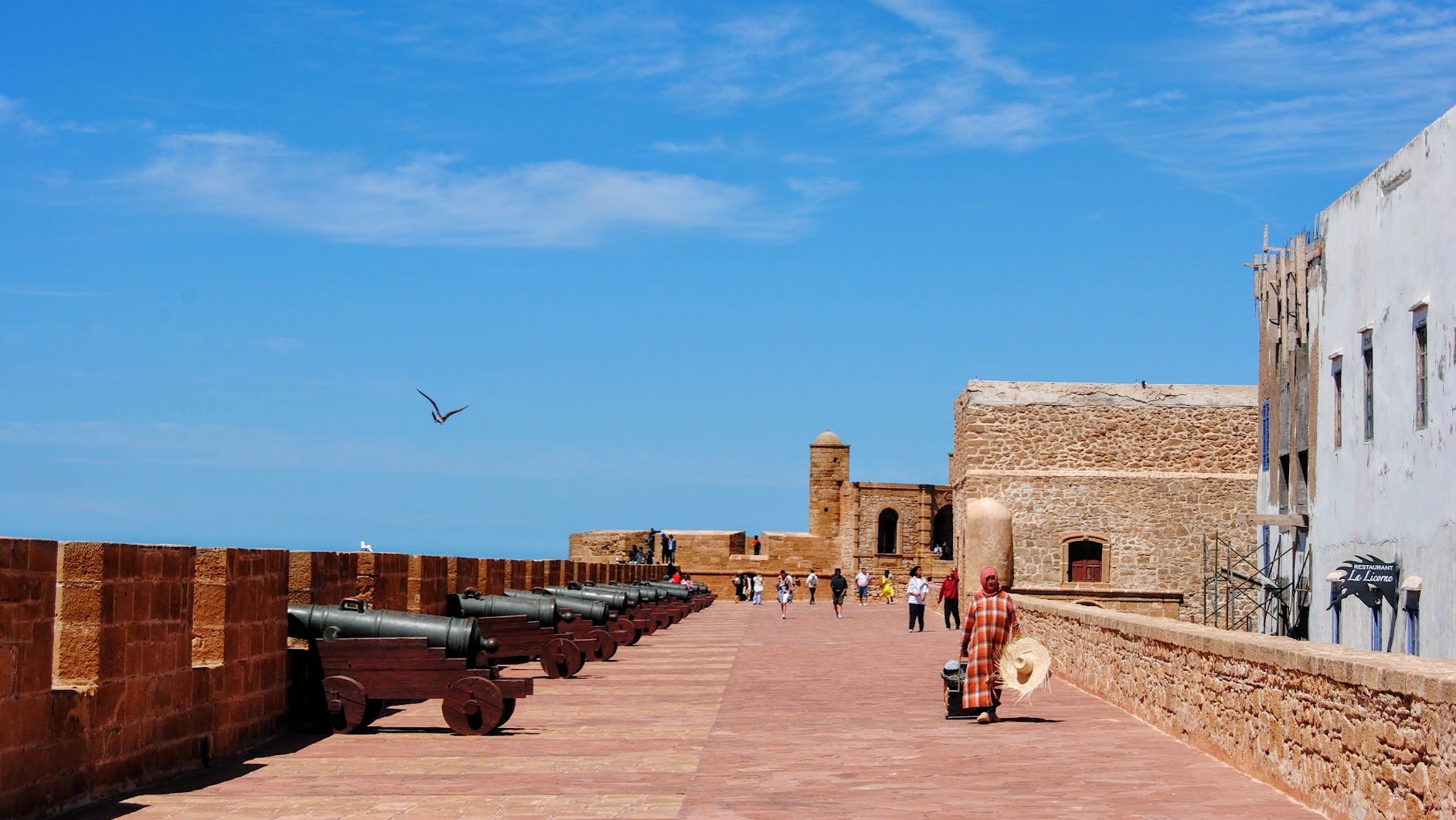 A woman carrying groceries and a large stray hat walks along the Essaouira city ramparts on a sunny day 