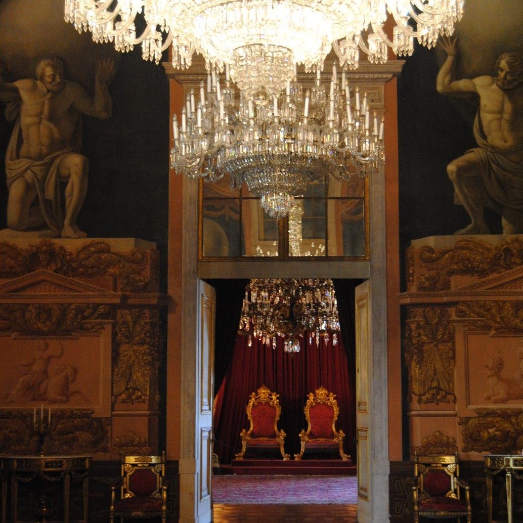A royal throne room with crystal chandeliers, murals, and art covering the walls.