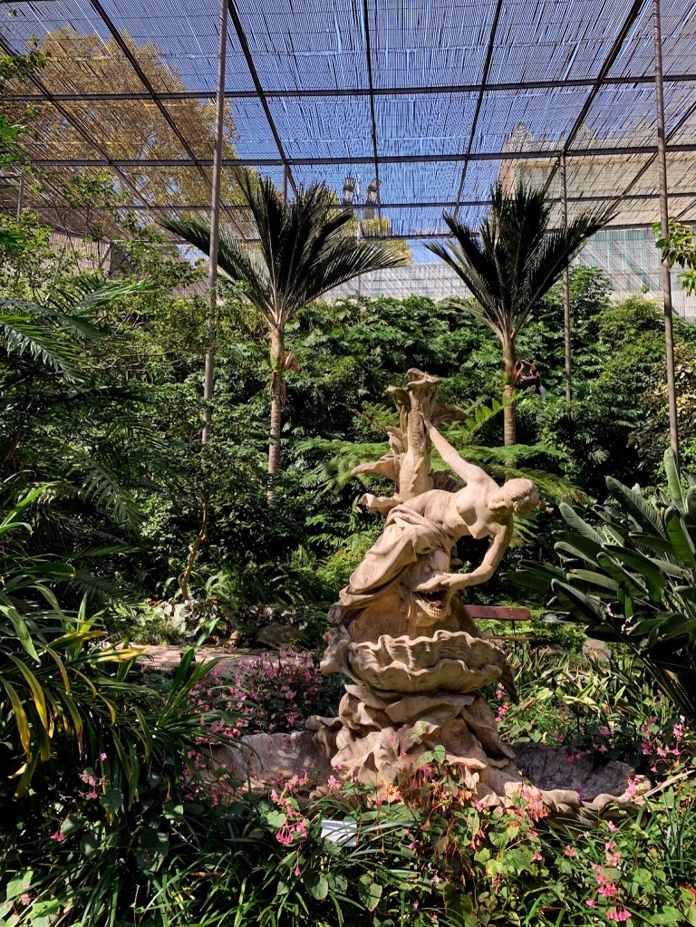 A statue of a woman in a large greenhouse surrounded by trees and pink flowers