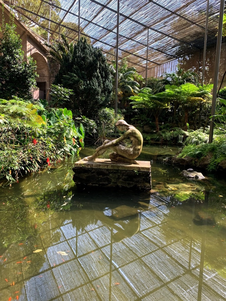 A statue of a woman sitting in the middle of a small pond in a greenhouse