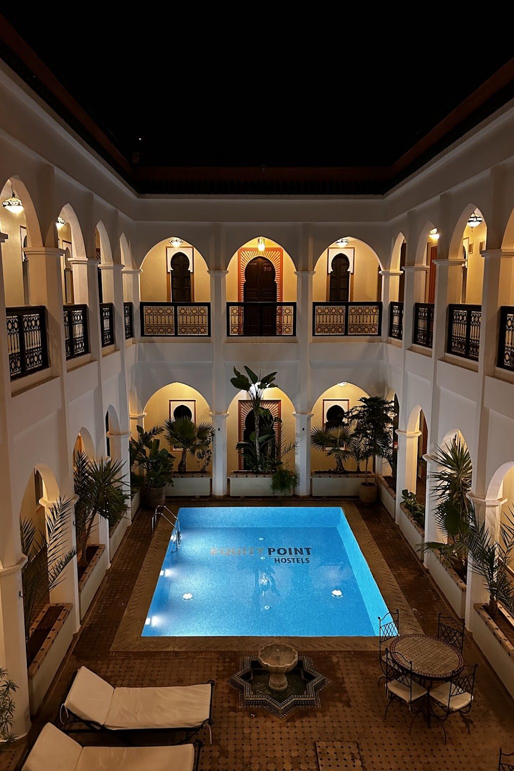 An empty riad courtyard at night with a pool, a table, decorative fountain, and lounge chairs.